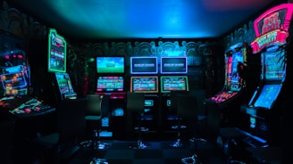 Dimly lit gaming arcade featuring a variety of brightly illuminated slot machines and video gaming screens. Neon signs and colorful displays create a vibrant atmosphere. The arrangement of gaming machines suggests an inviting space for entertainment, with chairs positioned for players.