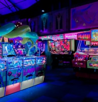 An indoor arcade filled with vibrant gaming machines lit with neon blue and pink lights. The scene includes various arcade games with flashing screens and colorful displays, and a large overhead structure resembling a green snake. The background shows illuminated signs and themed decor, contributing to an immersive entertainment atmosphere.