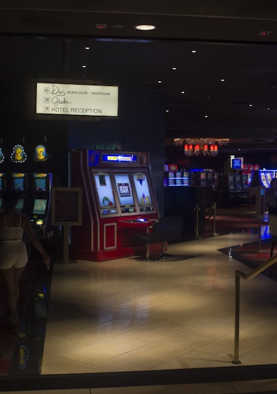 A dimly lit casino with several rows of slot machines, some displaying bright graphics and lights. There are a few people scattered throughout the area, engaging with the machines. The floor is reflective and polished, adding to the ambiance, while the ceiling is dotted with numerous small lights.