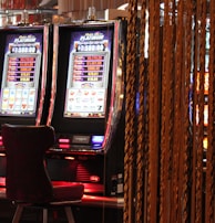 A pair of casino slot machines, each with detailed screens displaying various game outcomes and jackpot amounts. The chairs in front of the slot machines are dark and cushioned for comfort. The background features a hanging curtain made of golden ropes and other casino decorations are visible.
