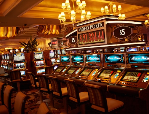 A casino floor featuring rows of brightly lit video poker machines. The area is illuminated by elegant chandeliers, with a display board showing a large sum of money, suggesting a possible jackpot. The rich decor includes luxurious carpets and comfortable seating, adding to the opulent atmosphere.