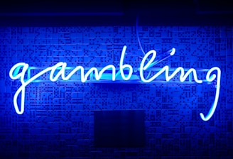 A bright blue neon sign spells out the word 'gambling' in cursive script. The background features a textured wall covered in domino tiles, also illuminated by the blue light, creating a vivid and visually striking scene.