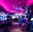 A vibrant arcade with rows of gaming machines adorned with colorful screens. Several people are immersed in playing games, seated on stools in a dimly lit room accented by an intense pink ceiling light. The atmosphere is lively and engaging.