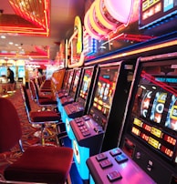 A brightly lit casino interior featuring a row of colorful slot machines. The neon lights create a vibrant atmosphere, with patterned carpeting adding to the decor. Stools are aligned neatly in front of each machine.
