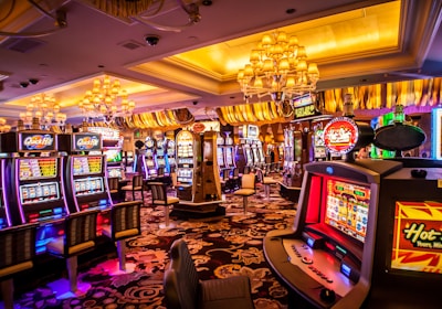 A vibrant casino interior filled with rows of colorful slot machines, each illuminated with bright lights and displays. The ceiling is adorned with elegant chandeliers and golden drapes, creating a luxurious atmosphere. The patterned carpet adds to the elaborate design of the space.