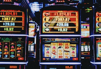 A row of colorful slot machines lit with bright neon lights, showcasing various graphical elements on the screens, indicating different games and jackpots. The atmosphere suggests a lively casino setting, bustling with excitement.