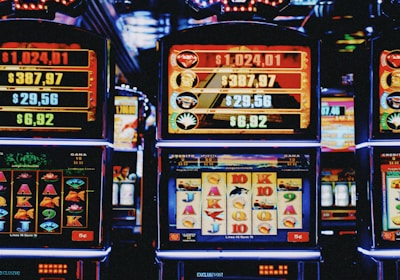 A row of colorful slot machines lit with bright neon lights, showcasing various graphical elements on the screens, indicating different games and jackpots. The atmosphere suggests a lively casino setting, bustling with excitement.
