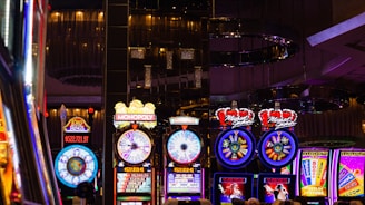 A lively casino scene featuring a row of bright, colorful slot machines with themes like Monopoly, Michael Jackson, and Lord of the Rings. The gaming machines display a variety of flashing lights and digital screens, creating an energetic and exciting atmosphere. Reflections of the lights are visible in glossy surfaces above the machines.