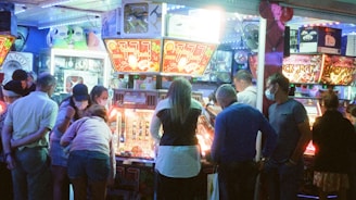 A group of people standing in front of an illuminated amusement arcade, engaging with various gaming machines. The environment is bustling with activity, and colorful lights create a vibrant atmosphere. The ceiling is decorated with large, playful images of dice and other game-related graphics, enhancing the lively mood.