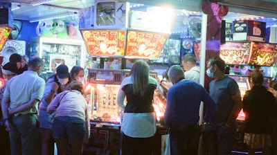 A group of people standing in front of an illuminated amusement arcade, engaging with various gaming machines. The environment is bustling with activity, and colorful lights create a vibrant atmosphere. The ceiling is decorated with large, playful images of dice and other game-related graphics, enhancing the lively mood.