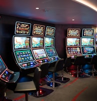 A row of brightly lit slot machines in a casino setting, each with colorful displays and various game themes. The machines are lined up against a dark wall and are unoccupied, with cushioned chairs placed in front of them. The area is dimly lit except for the glow from the screens, creating a vibrant and engaging atmosphere.