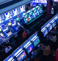 A bustling arcade filled with rows of gaming machines, each with vibrant displays and controls. Several people are seated at these machines, deeply engaged in playing games. Bright fluorescent lights cast a blue and neon glow across the room, enhancing the lively and energetic atmosphere.