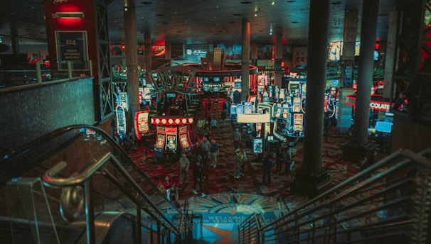 A bustling casino floor filled with groups of slot machines and people walking around. The atmosphere is vibrant with neon lights and illuminated signs. The interior has a ceiling supported by large pillars and there are stairs leading down to the gaming area.