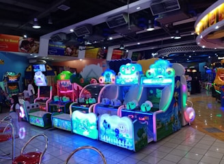 A brightly lit arcade filled with colorful gaming machines sits under ceiling lights. Neon lights illuminate various game stations, creating a playful and vibrant environment. A few red chairs are scattered around the reflective, tiled floor, and a person is observing one of the machines. The atmosphere is lively and cheerful, featuring games with cartoonish designs and engaging themes.