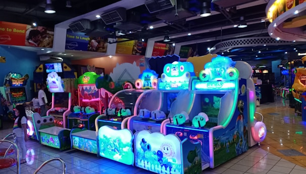 A brightly lit arcade filled with colorful gaming machines sits under ceiling lights. Neon lights illuminate various game stations, creating a playful and vibrant environment. A few red chairs are scattered around the reflective, tiled floor, and a person is observing one of the machines. The atmosphere is lively and cheerful, featuring games with cartoonish designs and engaging themes.